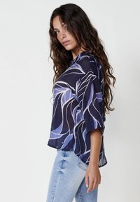 Blusa a maniche corte in tessuto leggero, con un motivo astratto blu scuro e viola chiaro, vestibilità rilassata, scollo rotondo, spacchetti laterali e orlo curvo.