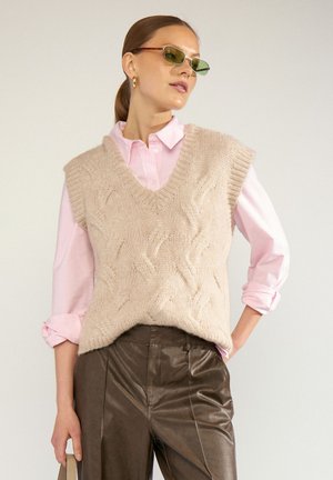 Femme aux cheveux attachés, portant des lunettes de soleil vertes, un gilet en tricot beige sur une chemise rose et un pantalon en cuir marron, posant avec une main dans la poche.