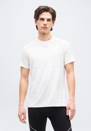 Mladi moški stoji, oblečen v belo kratkoročno atletsko majico in črne pajkice na svetlo sivi podlagi.