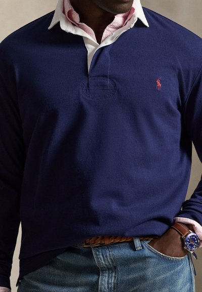 Man draagt een marineblauwe lange mouw polo over een roze overhemd met kraag, blauwe jeans, een bruine gevlochten riem en een blauw horloge.