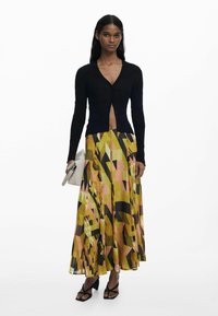 Cardigan noir à côtes avec un col en V et une fermeture à boutons, associé à une jupe à motifs vibrants jaune, rose et noir, portée avec des sandales noires.