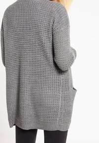 Szary sweter kardigan z dzianiny o otwartym splocie, z długimi rękawami i kieszenią po każdej stronie, sięgający do połowy uda.