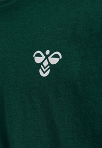 Białe stylizowane logo pszczoły na tle ciemnozielonej dzianiny.