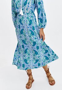 Długa niebiesko-zielona sukienka w wzór paisley, wykonana z lekkiego materiału, z balonowymi rękawami i rozkloszowanym dołem. Brązowe sandały na paski.
