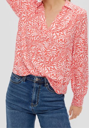 Dlouhý rukáv korálově bílé blůzy s vzorováním spárovaný s modrými džínami na osobě s jednou zvednutou rukou.