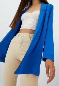 Blazer azul brillante de manga larga, combinado con una camiseta corta texturizada blanca y jeans ajustados beige claros. Diseño limpio, con detalles mínimos.