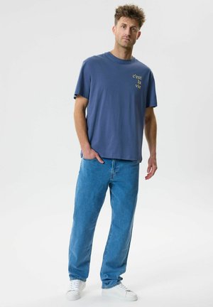 Mand stående med den ene hånd i lommen iført blå 'c'est la vie' t-shirt, lyseblå jeans og hvide sneakers på ensfarvet baggrund.