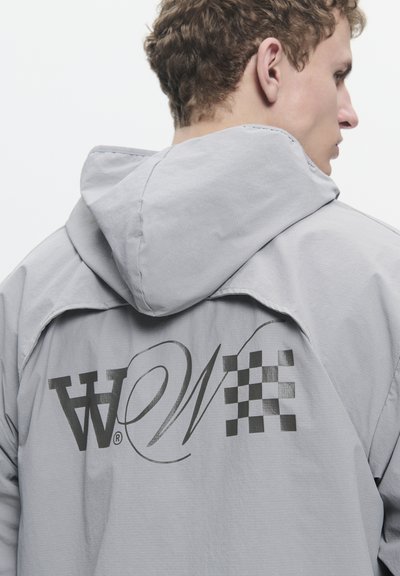 Veste à capuche gris clair avec un grand logo noir et un motif en damier au dos. Texture lisse avec une coupe décontractée.