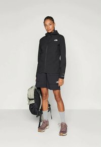 Must musta vihmane jakk, lukuga, kapuutsiga ja logoga. Kombineeritud tumedate lühikesed püksid, hallid sokid ja pruunid matkingingad. Kerge seljakott tumedate aktsentidega.