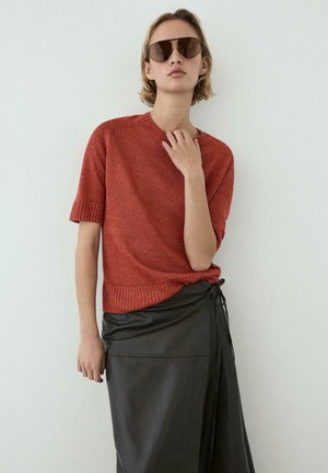 Kobieta w oversize brązowych okularach przeciwsłonecznych, rdzawej krótkiej dzianinowej bluzce i czarnej skórzanej spódnicy midi na jednolitym tle.