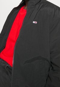Giacca leggera nera con chiusura a zip, dotata di colletto alto e piccolo logo ricamato. Indossata sopra una maglietta rossa.