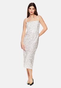 Silberne Paillettenkleid mit verstellbaren Spaghettiträgern, figurbetonter Silhouette, knielang, glatte Textur, getragen mit schwarzen spitzen Schuhen.