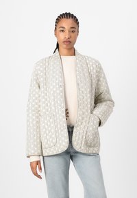 Chaqueta acolchada en crema con textura de patrón lineal, bolsillos profundos, apertura frontal y corte relajado, combinada con un suéter ligero y vaqueros.