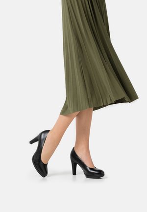Femme portant des chaussures à talons hauts noires et une jupe midi plissée olive, marchant avec un pied légèrement relevé.