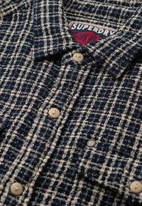 Navyblauwe en witte geruite wollen jas met beige knopen en een zichtbaar rood-wit "Superdry Work Wear" label binnen de kraag.
