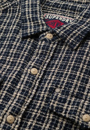 Chaqueta de lana a cuadros en azul marino y blanco con botones beige y una etiqueta visible en rojo y blanco de "Superdry Work Wear" dentro del cuello.