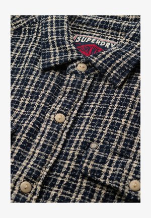 Giacca di lana a quadri blu navy e bianca con bottoni beigel e un'etichetta rossa e bianca "Superdry Work Wear" visibile all'interno del colletto.