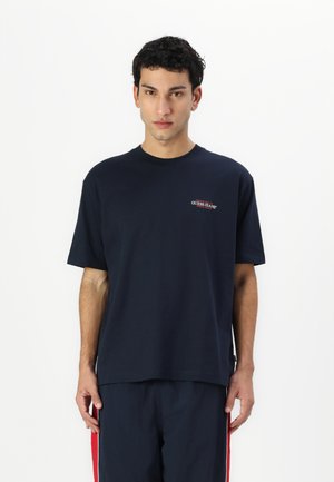 Maglietta in cotone blu navy con maniche corte, scollatura a girocollo e piccolo logo rosso "GUESS JEANS" sul petto a sinistra. Tessuto morbido, vestibilità comoda.