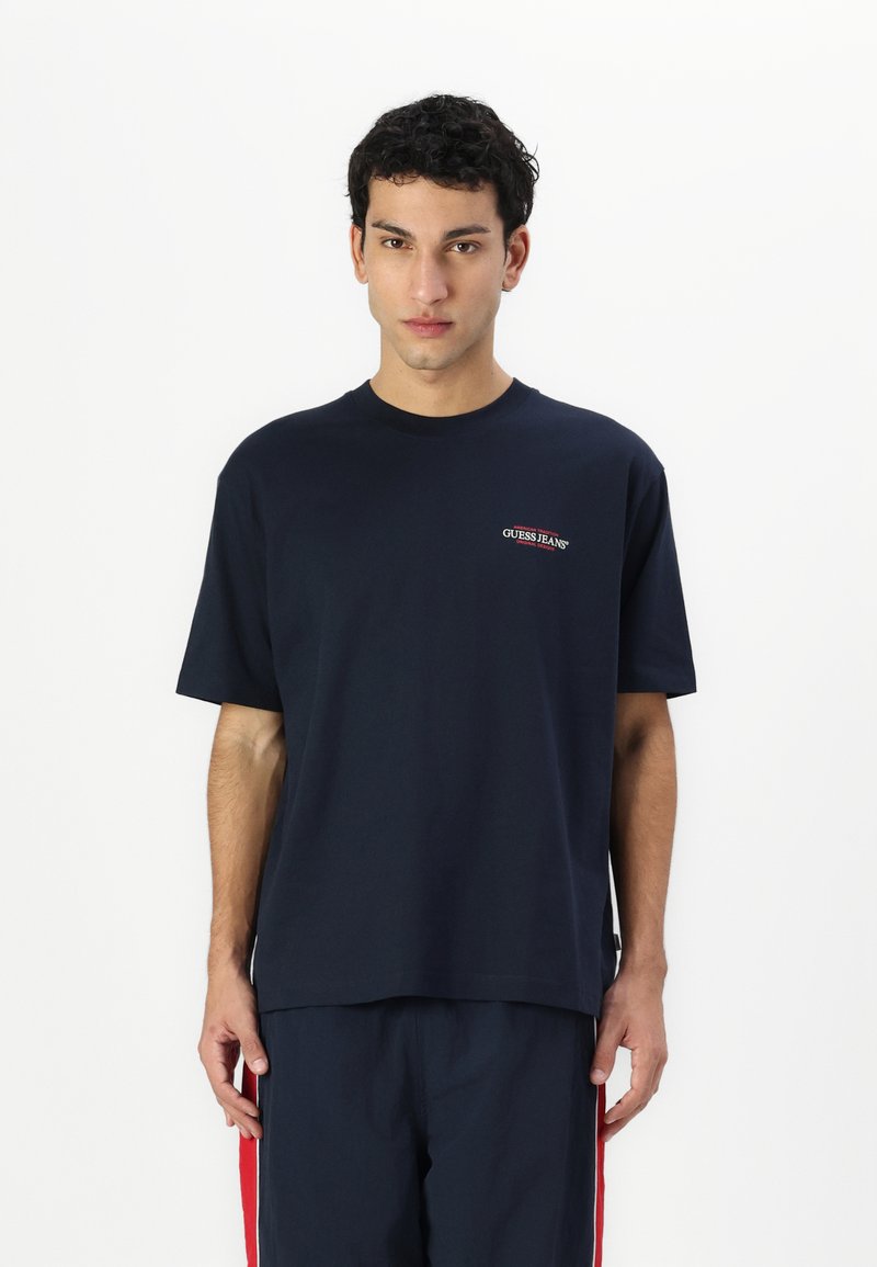 Sötétkék pamut póló rövid ujjakkal, kerek nyakkivágással és egy kis piros "GUESS JEANS" logóval a bal mellrészen. Sima tapintású, laza szabású.