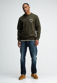 Olivengroene sweatshirt met een ronde hals en logo-detail op de linkerborst, gecombineerd met een blauwe spijkerbroek en tan kleur moccasin-schoenen.