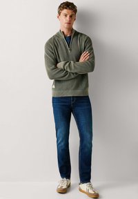 Jonge man staat met armen over elkaar, draagt een olijfgroene geribde trui, blauwe spijkerbroek en witte sneakers tegen een effen lichte achtergrond.