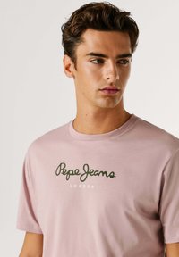 Roza bombažna T-majica z zelenim logotipom, ki na sprednji strani nosi napis "Pepe Jeans LONDON". Ima kratke rokave in okroglo ovratnico.