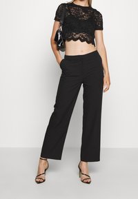 Top corto de encaje negro con dobladillo de pico, combinado con pantalones negros de corte ajustado. La modelo lleva tacones negros con tiras y lleva un bolso pequeño.