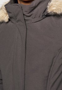 Mörkgrå vinterjacka med hög krage och beige syntetpäls på huvan, visas i närbild på övre bröst- och nackområde.