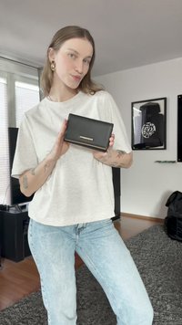 Mladá žena s tetováním v bílém tričku a džínech drží černou peněženku na úrovni hrudníku, stojí v moderním obývacím pokoji.