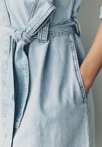 Ljust blå denimklänning med bälte i midjan, med framknappar, sidfickor och en lätt texturerad yta.