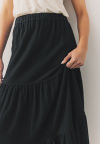 Czarna spódnica warstwowa wykonana z teksturowanego materiału, z marszczoną gumką w talii i subtelnymi pionowymi liniami. Ręka chwytająca materiał na dole.