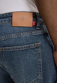 Niebieskie dżinsy z kontrastowym przeszyciem, brązowa skórzana naszywka w pasie z tłoczonym tekstem oraz czerwona metka na szwie bocznym.