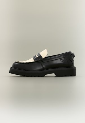 Schwarz-weißer Lederloafer mit schwarzer dicksohliger Sohle und Nahtdetail, von der Außenseite auf einfarbigem Hintergrund gesehen.