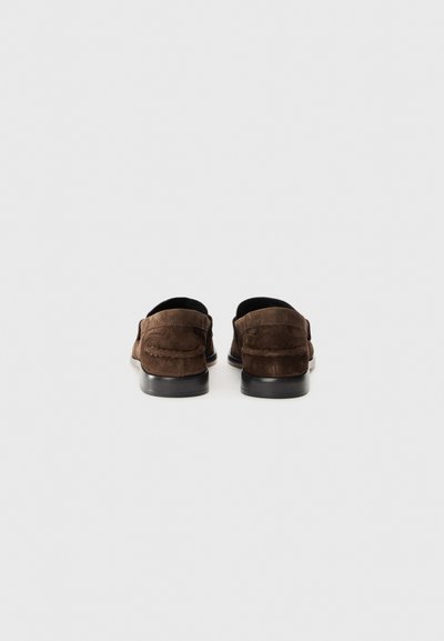 Mocasines de ante marrón con una textura suave, forma redondeada y suelas oscuras en contraste. Presenta costuras visibles a lo largo de la parte posterior.