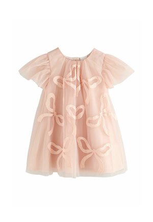 Mladé dievča v svetloružových šatách s motívmi mašlí a trblietave ružové topánky Mary Jane, stojace proti neutrálneho pozadia.