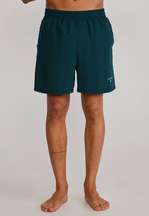 Homme portant un short vert foncé à taille élastique, debout pieds nus contre un fond uni, le short arbore un petit logo blanc sur la jambe gauche.