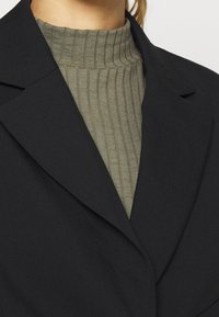 Veste noire à col à revers portée sur un col roulé côtelé vert olive. Le tissu semble lisse avec une coupe structurée.