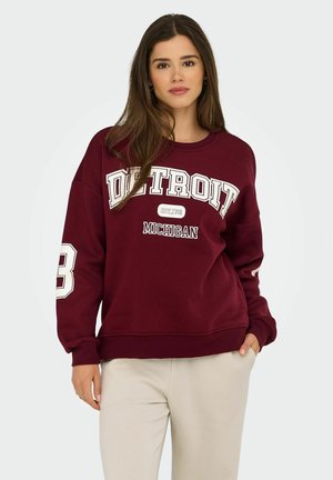 Burgundy sweatshirt featuring "DETROIT" in white text, an oversized fit, ribbed cuffs, and a round neckline. Paired with light beige pants.