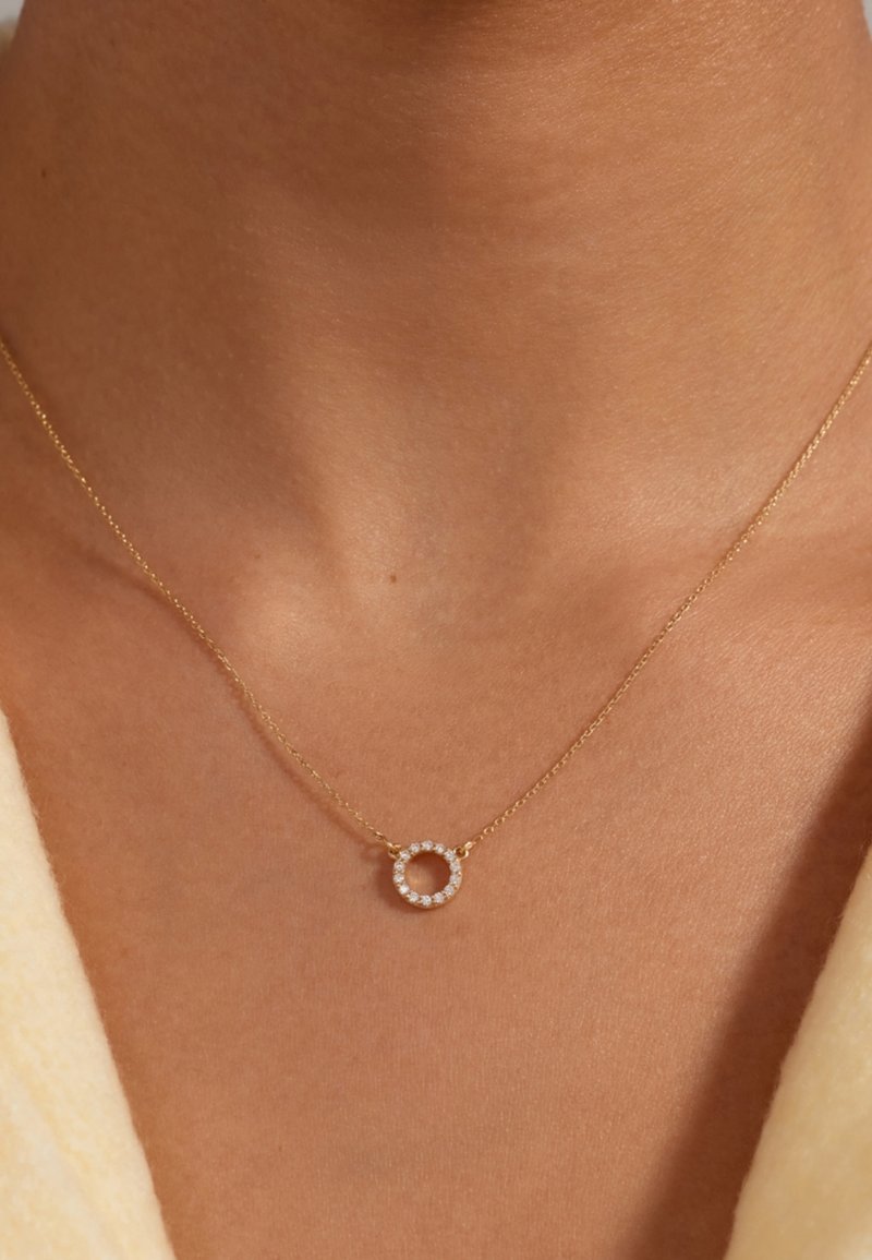 Collier en chaîne dorée avec un pendentif circulaire orné de petites pierres transparentes, suspendu élégamment contre un teint de peau neutre.