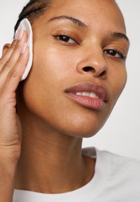 Close-up på en person som applicerar en bomullspad på sin kind. Huden verkar slät, medelhavston; bär en enkel vit skjorta.