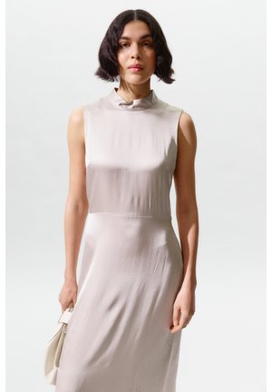 Nő rövid, hullámos, sötét hajjal, világosszürke ujjatlan ruhában, kezében egy kis, hozzáillő kézitáskával, egyszínű háttér előtt áll.