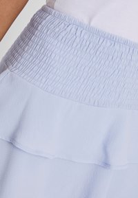 Gonna azzurro chiaro con vita smock e design a strati di rouches. Il materiale è morbido con una texture fluida e una leggera lucentezza.