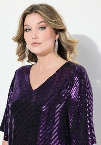 Haut en sequins violet à encolure en V avec des manches courtes, présentant une texture étincelante et une coupe ajustée, associé à de longues boucles d'oreilles argentées pendantes.