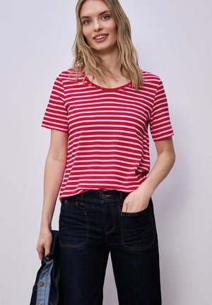 Jeune femme souriante, portant un t-shirt à manches courtes rayé rouge et blanc et un jean foncé, tenant une veste en jean dans une main.