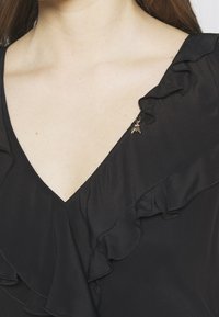 La blusa nera presenta una scollatura a V con dettagli di rouches lungo i bordi. Un piccolo accento di ape metallica si trova sulla spalla.