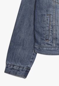 Rukáv džínové bundy v tmavě modré barvě s klasickým střihem. Má kapsu a prošité manžety. Hladká textura s viditelnými švy.