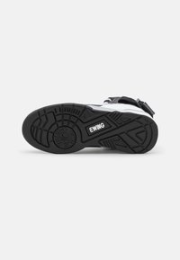 Semelle de chaussure de sport noire et blanche avec un motif de rainures géométriques et le logo "EWING", présentée sur un fond blanc uni.