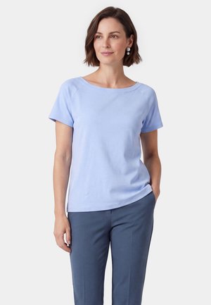 Donna con capelli corti castani, indossa una maglietta azzurro chiaro a maniche corte e pantaloni blu scuro, in piedi con una mano in tasca.