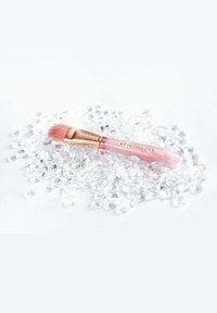 Pędzel do makijażu z różowym uchwytem i złotymi akcentami, z miękkimi różowymi włoskami, umieszczony na poduszce z przezroczystych ozdobnych kamieni.