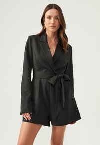 Black satin blazer dress featuring long sleeves, a deep V-neck, a tied waist belt, and tailored shorts. Smooth texture with minimal detailing.